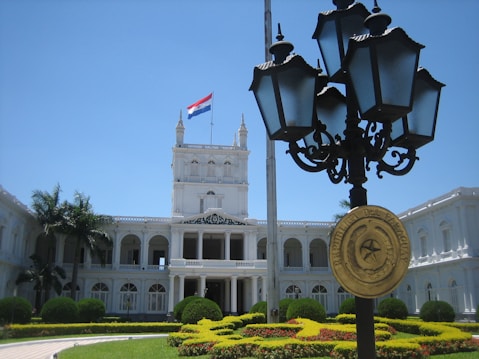 A majestic, white neoclassical building with multiple arches and columns, surrounded by lush greenery and well-maintained gardens. A flag featuring red, white, and blue stripes is flying atop the building. In the foreground, a decorative street lamp with a plaque reading 'Republica del Paraguay' is prominently displayed.