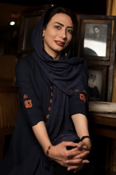 A woman with a gentle smile is seated, wearing a dark navy blue headscarf and dress adorned with intricate embroidery on the sleeves. She is sitting with her hands clasped in front of her, against a backdrop that includes framed vintage photographs. The setting is dimly lit with a warm ambiance.