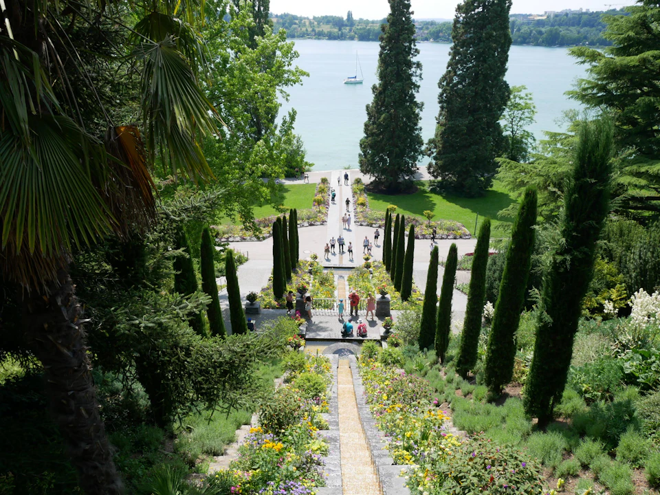 The image depicts a serene garden scene with a long stone staircase leading down to a body of water, possibly a lake or river. The stairs are adorned with an array of colorful flowers and plants, adding vibrancy to the landscape. Several people can be seen walking along the path on either side of the stairs, enjoying the tranquil surroundings. In the distance, there is a boat floating on the water, further enhancing the picturesque setting.