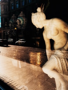 A white marble statue of a woman is prominently displayed, with light casting dramatic shadows and reflections through a glass window. Behind the statue are ornate vases with intricate designs, and the exterior reflects an urban street scene with buildings and vehicles.