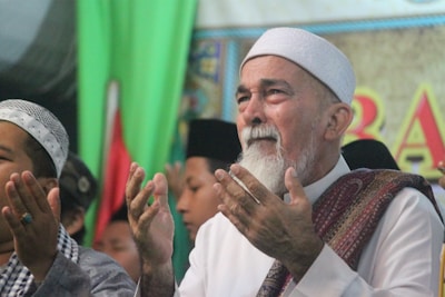 An elderly man with a white beard and wearing a white headscarf stands with his hands raised in a prayer gesture. He appears emotional; his eyes are closed and he has a solemn expression on his face. Surrounding him are other men, some of whom are also engaged in prayer. The background is decorated with a green and colorful banner.