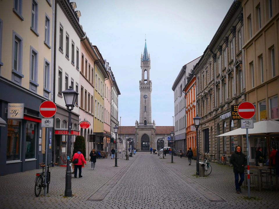 The image depicts a narrow cobblestone street flanked by buildings on both sides, with a tall clock tower standing out against the sky. The street is bustling with activity, featuring several people walking and biking along its length. On one side of the street, there are two red traffic signs - a "do not enter" sign and a "no parking" sign. In the background, a church can be seen, adding to the historical charm of the scene.