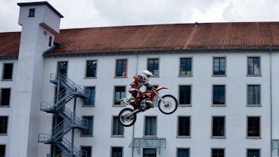 A motorcyclist is performing a high jump on a dirt bike in front of a large building with multiple windows and a red-tiled roof. The motorcycle is airborne, demonstrating a dynamic stunt scene.