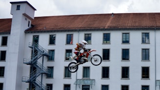 A motorcyclist is performing a high jump on a dirt bike in front of a large building with multiple windows and a red-tiled roof. The motorcycle is airborne, demonstrating a dynamic stunt scene.