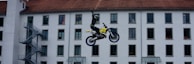 A motorcyclist is performing a mid-air stunt in front of a multi-story building with numerous windows. The rider is raising one hand while balancing on a yellow and black dirt bike. The building facade has a red-tiled roof and several windows are open, with some people visible looking out.