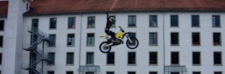 A motorcyclist is performing a mid-air stunt in front of a multi-story building with numerous windows. The rider is raising one hand while balancing on a yellow and black dirt bike. The building facade has a red-tiled roof and several windows are open, with some people visible looking out.