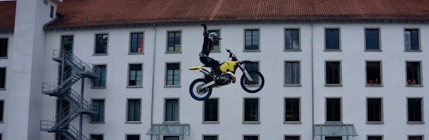 A motorcyclist is performing a mid-air stunt in front of a multi-story building with numerous windows. The rider is raising one hand while balancing on a yellow and black dirt bike. The building facade has a red-tiled roof and several windows are open, with some people visible looking out.