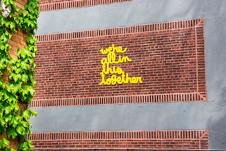 A brick wall with a handwritten message in bright yellow text reads 'we're all in this together.' The wall is partially covered with vibrant green ivy leaves on the left side.