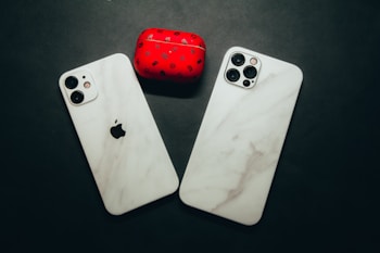 Two smartphones with marble-patterned cases are lying on a dark surface next to a red case with black designs. The phones have a minimal and sleek design with visible camera lenses.