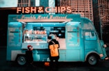 A vintage-style food truck painted in bright turquoise serves fish and chips, adorned with a marquee sign lights. It is located in an urban setting with tall buildings in the background. A customer with a yellow backpack and a child in a red hat stand at the counter.