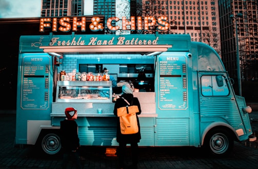 A vintage-style food truck painted in bright turquoise serves fish and chips, adorned with a marquee sign lights. It is located in an urban setting with tall buildings in the background. A customer with a yellow backpack and a child in a red hat stand at the counter.