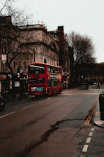 A classic red double-decker bus winding through a historic London street lined with old stone buildings.