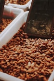 A large container filled with almonds, with a dark, slightly out-of-focus background that includes a chalkboard with partially visible writing.
