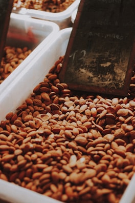 A large container filled with almonds, with a dark, slightly out-of-focus background that includes a chalkboard with partially visible writing.