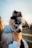 A small, fluffy puppy with striking blue eyes is being held gently. The dog has a distinctive black and white coat with tan markings and wears a blue and white checkered bandana. The image is softly lit, suggesting an outdoor setting, possibly during a sunset.