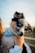 A small, fluffy puppy with striking blue eyes is being held gently. The dog has a distinctive black and white coat with tan markings and wears a blue and white checkered bandana. The image is softly lit, suggesting an outdoor setting, possibly during a sunset.