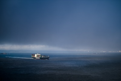 A large container ship travels across the ocean under a dark, overcast sky. The ship, loaded with colorful containers, creates a trail in the water as it moves through the deep blue sea. A faint horizon is visible in the background, suggesting distant land or structures.