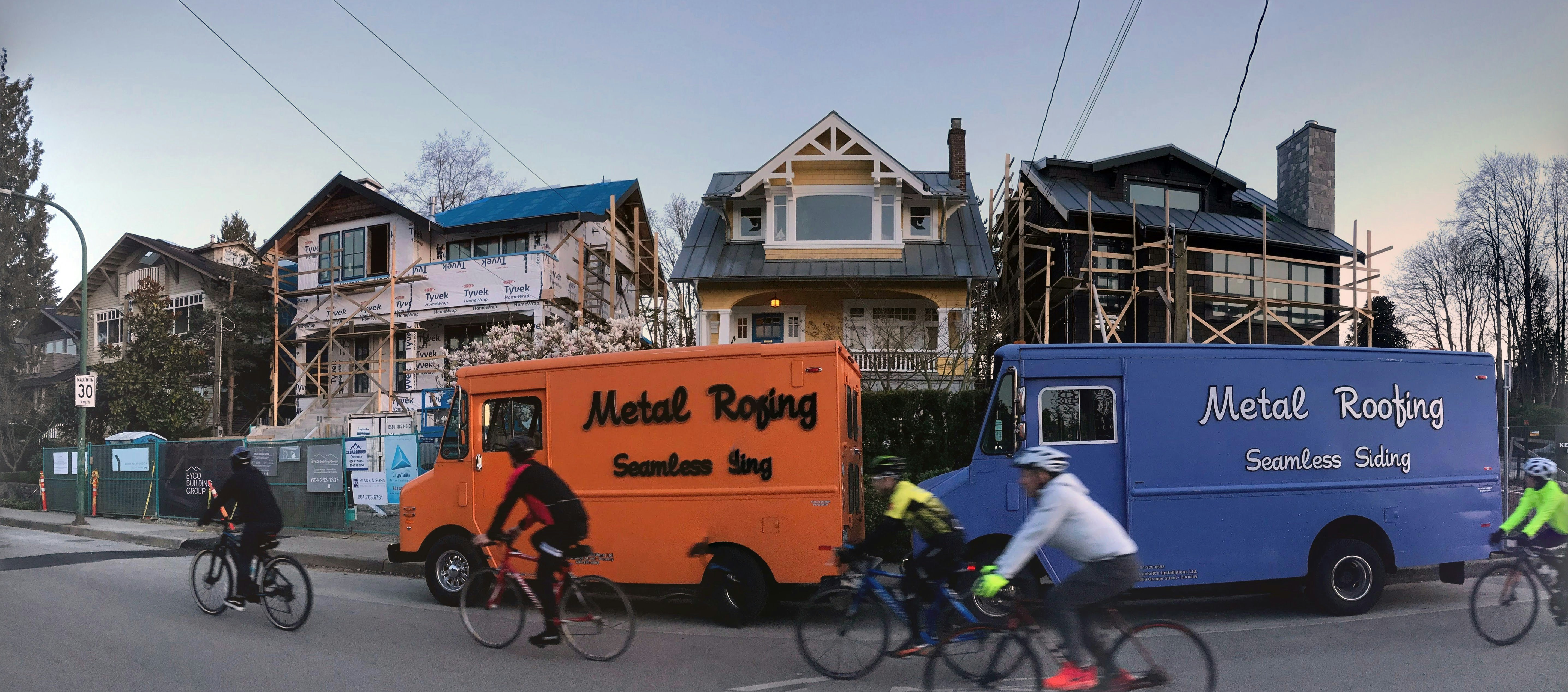 A residential street with three houses in the background, two of which are under construction or renovation. Two vans labeled 'Metal Roofing Seamless Siding' are parked on the side of the street, one orange and one blue. Several cyclists are riding past, wearing helmets and cycling gear.
