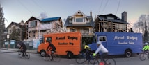 A residential street with three houses in the background, two of which are under construction or renovation. Two vans labeled 'Metal Roofing Seamless Siding' are parked on the side of the street, one orange and one blue. Several cyclists are riding past, wearing helmets and cycling gear.