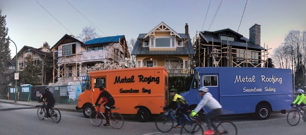 A residential street with three houses in the background, two of which are under construction or renovation. Two vans labeled 'Metal Roofing Seamless Siding' are parked on the side of the street, one orange and one blue. Several cyclists are riding past, wearing helmets and cycling gear.