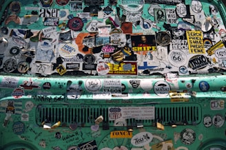 A turquoise vehicle's surface is covered in various colorful and intricate stickers, including travel logos, brand names, artistic designs, and textual slogans. The stickers overlap and are of different shapes and sizes, creating a chaotic yet vibrant patchwork. Some of the text includes road names and artistic expressions.