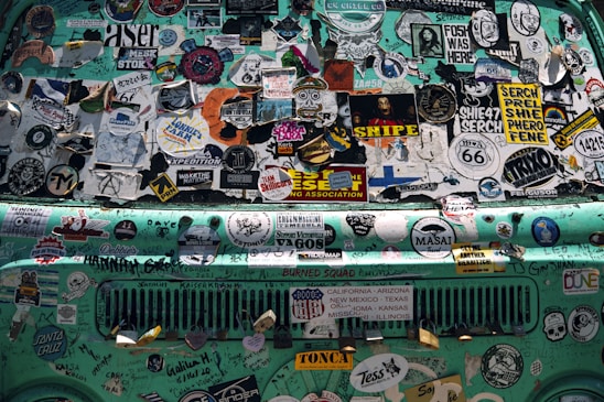 A turquoise vehicle's surface is covered in various colorful and intricate stickers, including travel logos, brand names, artistic designs, and textual slogans. The stickers overlap and are of different shapes and sizes, creating a chaotic yet vibrant patchwork. Some of the text includes road names and artistic expressions.