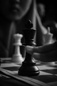 A close-up of a chess game in progress, showing a hand moving a black king piece on the board. The background is blurred, and there is a white queen piece visible to the side. The composition focuses on the intense concentration and the strategic moment in the game.