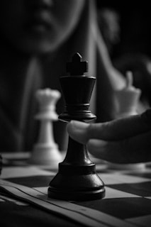 A close-up of a chess game in progress, showing a hand moving a black king piece on the board. The background is blurred, and there is a white queen piece visible to the side. The composition focuses on the intense concentration and the strategic moment in the game.