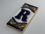 A decorative ceramic tile featuring a large, dark blue letter 'R' centered on a white background. The tile is framed with a colorful mosaic pattern, including green, red, yellow, and blue triangles and rectangles along the top and bottom edges.