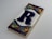 A decorative ceramic tile featuring a large, dark blue letter 'R' centered on a white background. The tile is framed with a colorful mosaic pattern, including green, red, yellow, and blue triangles and rectangles along the top and bottom edges.