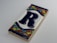 A decorative ceramic tile featuring a large, dark blue letter 'R' centered on a white background. The tile is framed with a colorful mosaic pattern, including green, red, yellow, and blue triangles and rectangles along the top and bottom edges.