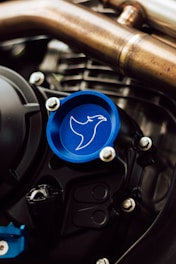 A close-up view of a mechanical component featuring a blue cap with an outline of a bird or hawk. The surroundings include metallic pipes and bolts, suggestive of an engine or motor part. The blue cap stands out against the metallic hues and complex structures.