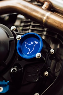 A close-up view of a mechanical component featuring a blue cap with an outline of a bird or hawk. The surroundings include metallic pipes and bolts, suggestive of an engine or motor part. The blue cap stands out against the metallic hues and complex structures.