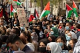 A large group of people are gathered in a public area, many wearing face masks. They are holding Palestinian flags and a sign that reads 'Free Palestine'. The crowd appears engaged, with several individuals taking photos and videos with their phones.