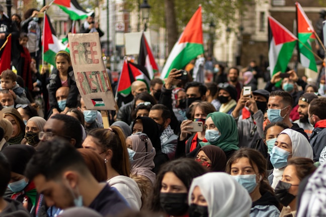 A large group of people are gathered in a public area, many wearing face masks. They are holding Palestinian flags and a sign that reads 'Free Palestine'. The crowd appears engaged, with several individuals taking photos and videos with their phones.