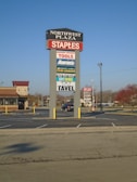 A tall sign located in a shopping plaza parking lot displays various business names such as Staples, Harbor Freight Tools, and Aaron's. The sign is set against a clear blue sky, with additional stores and trees visible in the background.