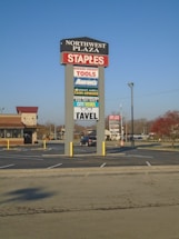 A tall sign located in a shopping plaza parking lot displays various business names such as Staples, Harbor Freight Tools, and Aaron's. The sign is set against a clear blue sky, with additional stores and trees visible in the background.