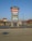 A tall sign located in a shopping plaza parking lot displays various business names such as Staples, Harbor Freight Tools, and Aaron's. The sign is set against a clear blue sky, with additional stores and trees visible in the background.