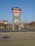 A tall sign located in a shopping plaza parking lot displays various business names such as Staples, Harbor Freight Tools, and Aaron's. The sign is set against a clear blue sky, with additional stores and trees visible in the background.