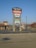 A tall sign located in a shopping plaza parking lot displays various business names such as Staples, Harbor Freight Tools, and Aaron's. The sign is set against a clear blue sky, with additional stores and trees visible in the background.