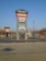 A tall sign located in a shopping plaza parking lot displays various business names such as Staples, Harbor Freight Tools, and Aaron's. The sign is set against a clear blue sky, with additional stores and trees visible in the background.