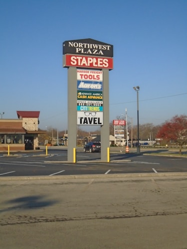 A tall sign located in a shopping plaza parking lot displays various business names such as Staples, Harbor Freight Tools, and Aaron's. The sign is set against a clear blue sky, with additional stores and trees visible in the background.