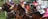 A group of jockeys riding racehorses are caught in mid-action, all wearing colorful racing silks and helmets. They appear to be at the starting gate, with focused and determined expressions as they begin a race. The background includes greenery and parts of the racing infrastructure.