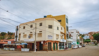 A street corner with a multi-story building featuring various signs and businesses on the ground floor. The building has a beige and mustard color scheme, with utility wires crisscrossing in the foreground. Several parked vehicles, including a motorcycle and a car, can be seen along the street. To the left, there are stalls with blue and white coverings, and the background shows a residential area on a hillside.