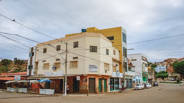A street corner with a multi-story building featuring various signs and businesses on the ground floor. The building has a beige and mustard color scheme, with utility wires crisscrossing in the foreground. Several parked vehicles, including a motorcycle and a car, can be seen along the street. To the left, there are stalls with blue and white coverings, and the background shows a residential area on a hillside.