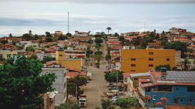 Several houses with brown and red tiled roofs stretch across a suburban area. The streets are lined with trees and utility poles, with a yellow and a blue building in the foreground. The landscape is slightly hilly with greenery and palms dotting the area.