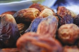 A close-up view of a pile of dried dates, showcasing their wrinkled texture and varied shades of brown and golden hues. The soft focus in the background emphasizes the rich, natural detail of the dates in the foreground.