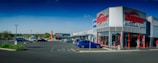 A commercial shopping area with a 'Tiger Wheel & Tyre' store prominently in the foreground, featuring large signage. Several other storefronts are visible in the background, including 'Tile Africa'. The parking lot has a few parked cars, and the weather appears clear with a deep blue sky.