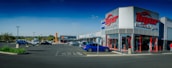 A commercial shopping area with a 'Tiger Wheel & Tyre' store prominently in the foreground, featuring large signage. Several other storefronts are visible in the background, including 'Tile Africa'. The parking lot has a few parked cars, and the weather appears clear with a deep blue sky.
