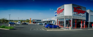 A commercial shopping area with a 'Tiger Wheel & Tyre' store prominently in the foreground, featuring large signage. Several other storefronts are visible in the background, including 'Tile Africa'. The parking lot has a few parked cars, and the weather appears clear with a deep blue sky.